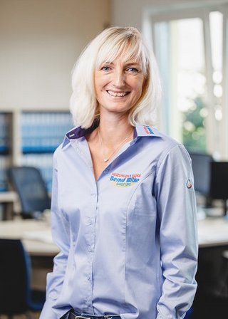 Die Frau mit blonden Haaren und einem freundlichen Lächeln trägt ein Hemd mit dem Logo eines Unternehmens in einem hellen Büro.