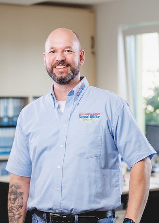 Der Mann mit tätowiertem Arm strahlt Freundlichkeit aus, während er in einem modernen Büro mit natürlichem Licht posiert.