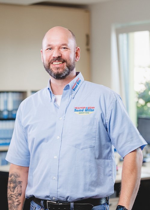 Der Mann mit tätowiertem Arm strahlt Freundlichkeit aus, während er in einem modernen Büro mit natürlichem Licht posiert.