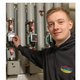 Ein junger Mann in einem schwarzen Hoodie steht vor Heizungsanlagen und überprüft einen digitalen Zähler mit einem Lächeln.