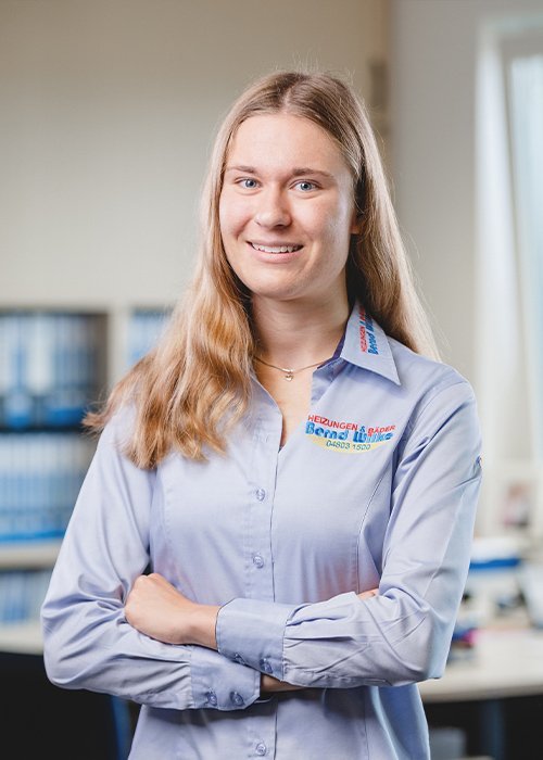 Die Person trägt ein hellblaues Hemd mit einem Logo und hat die Arme vor der Brust verschränkt, was Selbstbewusstsein ausstrahlt.