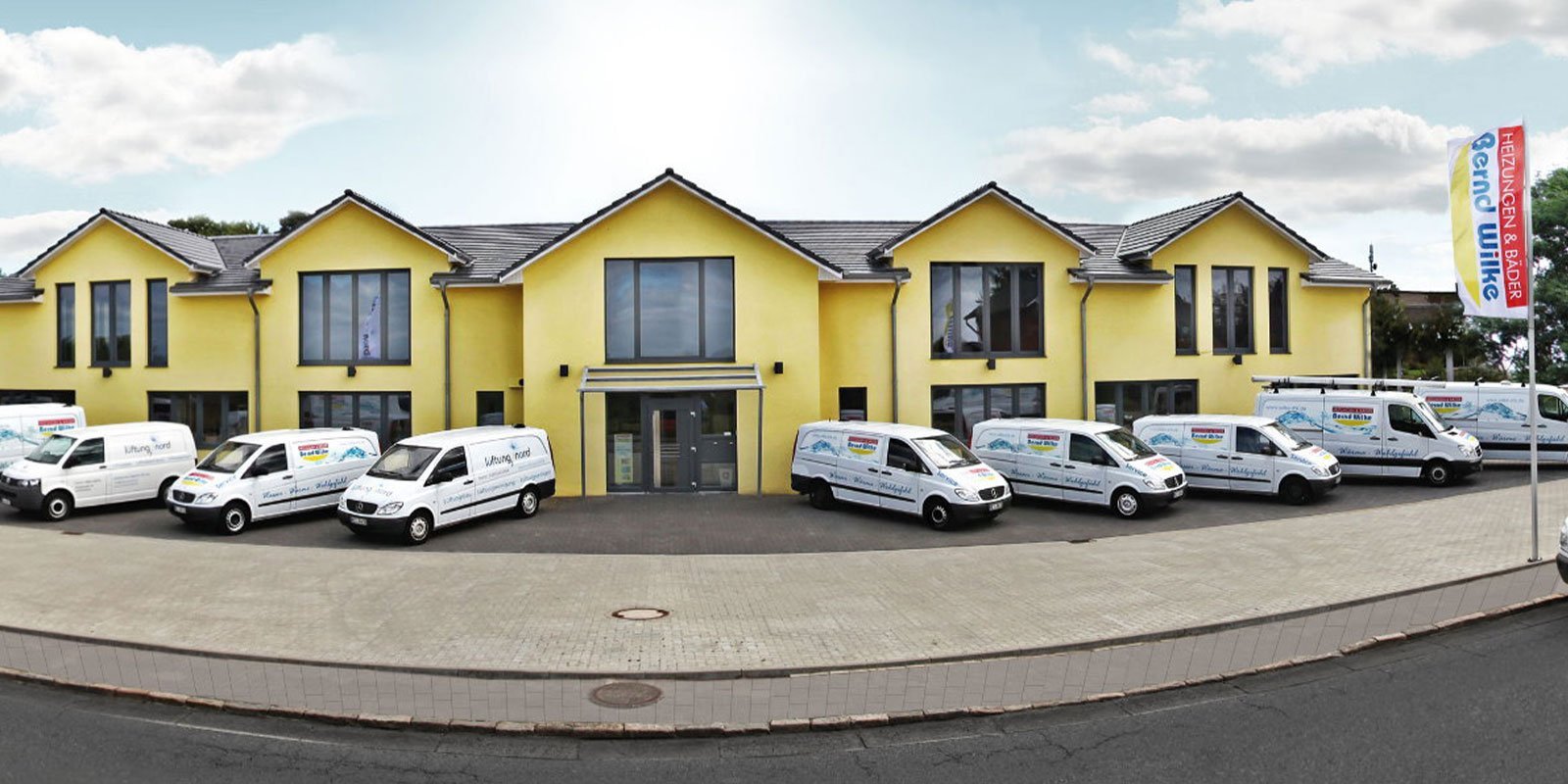 Das gelbe Firmengebäude mit einem einladenden Eingang, umgeben von mehreren weißen Lieferwagen, die auf einem gepflasterten Platz stehen.