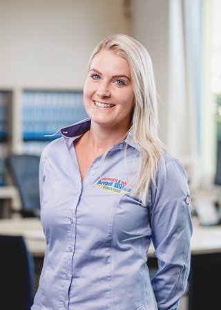 Die junge Frau steht in einem Büro mit Aktenregalen im Hintergrund und zeigt ein strahlendes Lächeln, das Vertrauen ausstrahlt.