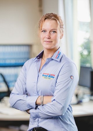Die Frau mit blonden Haaren trägt ein Hemd mit dem Logo eines Unternehmens und blickt direkt in die Kamera.