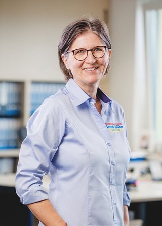Die Frau mit kurzen Haaren und Brille strahlt Freundlichkeit aus, während sie in einem gut beleuchteten Büro posiert.