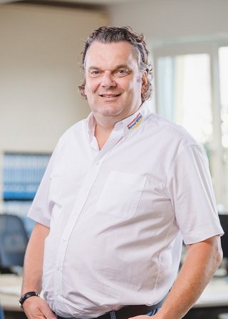 In einem hellen Büro steht ein lächelnder Mann in einem weißen Hemd, umgeben von Büromöbeln und einem freundlichen Ambiente.