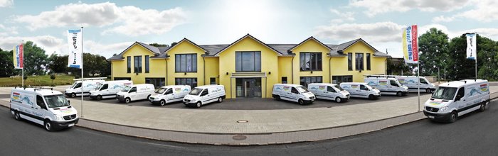 Vor einem modernen, gelben Firmengebäude stehen zahlreiche weiße Transporter, die mit dem Logo eines Heizungsunternehmens beschriftet sind.