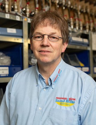 Der Mann mit Brille und einem blauen Hemd posiert vor Regalen, die mit verschiedenen Werkzeugen und Zubehör gefüllt sind.