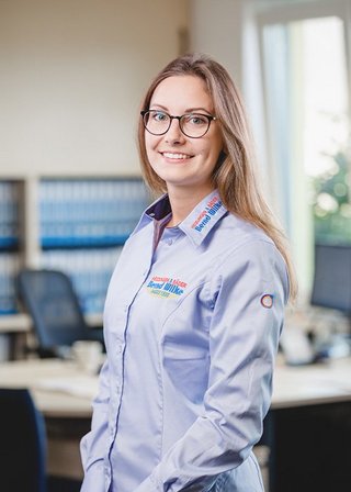 Die Frau mit langen, blonden Haaren steht in einem Büro und strahlt Selbstbewusstsein aus, während sie in die Kamera lächelt.