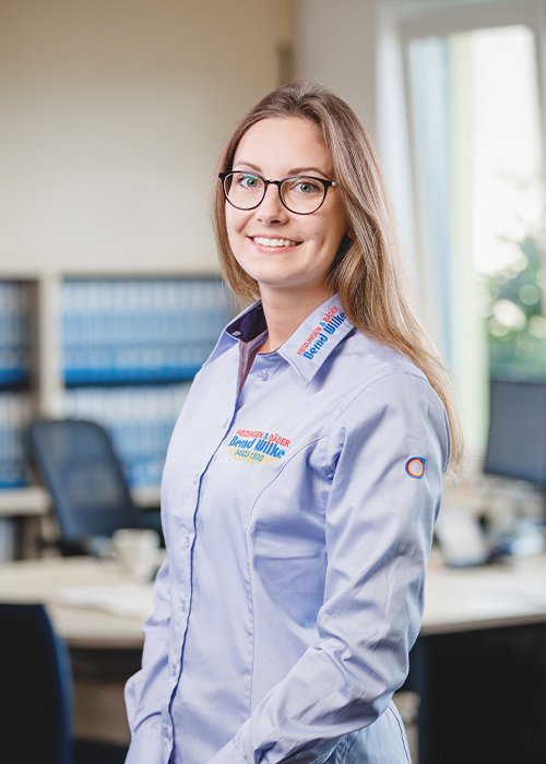 Die Frau mit langen, blonden Haaren steht in einem Büro und strahlt Selbstbewusstsein aus, während sie in die Kamera lächelt.