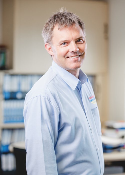 Der Mann mit kurzen, grauen Haaren zeigt ein freundliches Lächeln, während er in einem modernen Büro posiert.