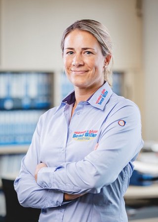 In einem hellen Büro steht eine Frau mit blonden Haaren, die ein professionelles Hemd trägt und eine positive Ausstrahlung hat.